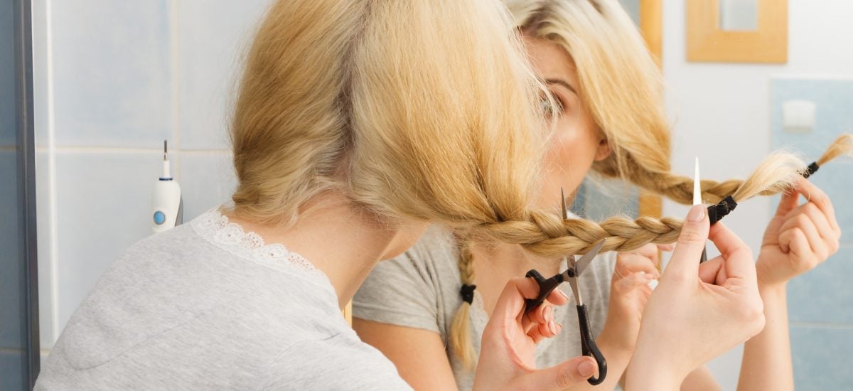 Žena strihajúca si nožničkami blond vlasy pred zrkadlom